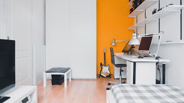 Ein Zimmer mit einem Schreibtisch, Fernseher, Kleiderschrank , Gitarre und einer Orangen Wandfarbe